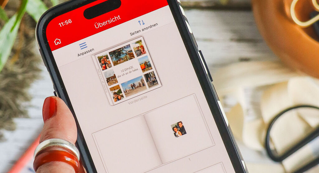 V ospredju slike ženska roka z rdeče nalakiranimi nohti drži pametni telefon. Na zaslonu je odprt vmesnik za urejanje mobilne aplikacije CEWE PHOTO. To je prej predstavljena fotoknjiga, ki jo je mogoče videti v zamegljenem ozadju slike na površini mize. Zamegljeno je mogoče videti tudi zeleno rastlino in ustvarjalne pripomočke, kot so pisani okrasni trakovi, pisala, škarje in darilni trak.